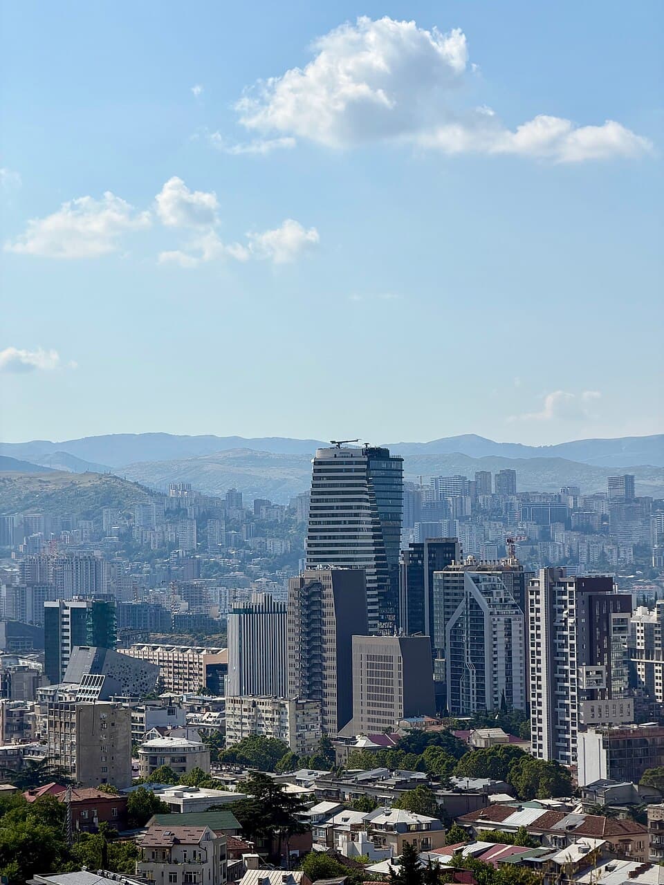 Modern city skyline with a prominent striped skyscraper and distant mountains under a cloudy sky.