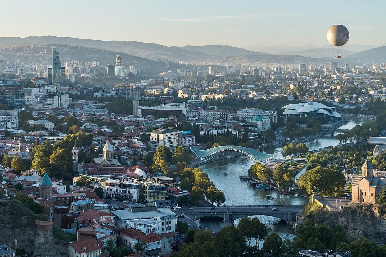 Aerial view of Tbilisi featuring Kura River, Bridge of Peace, and a hot air balloon.